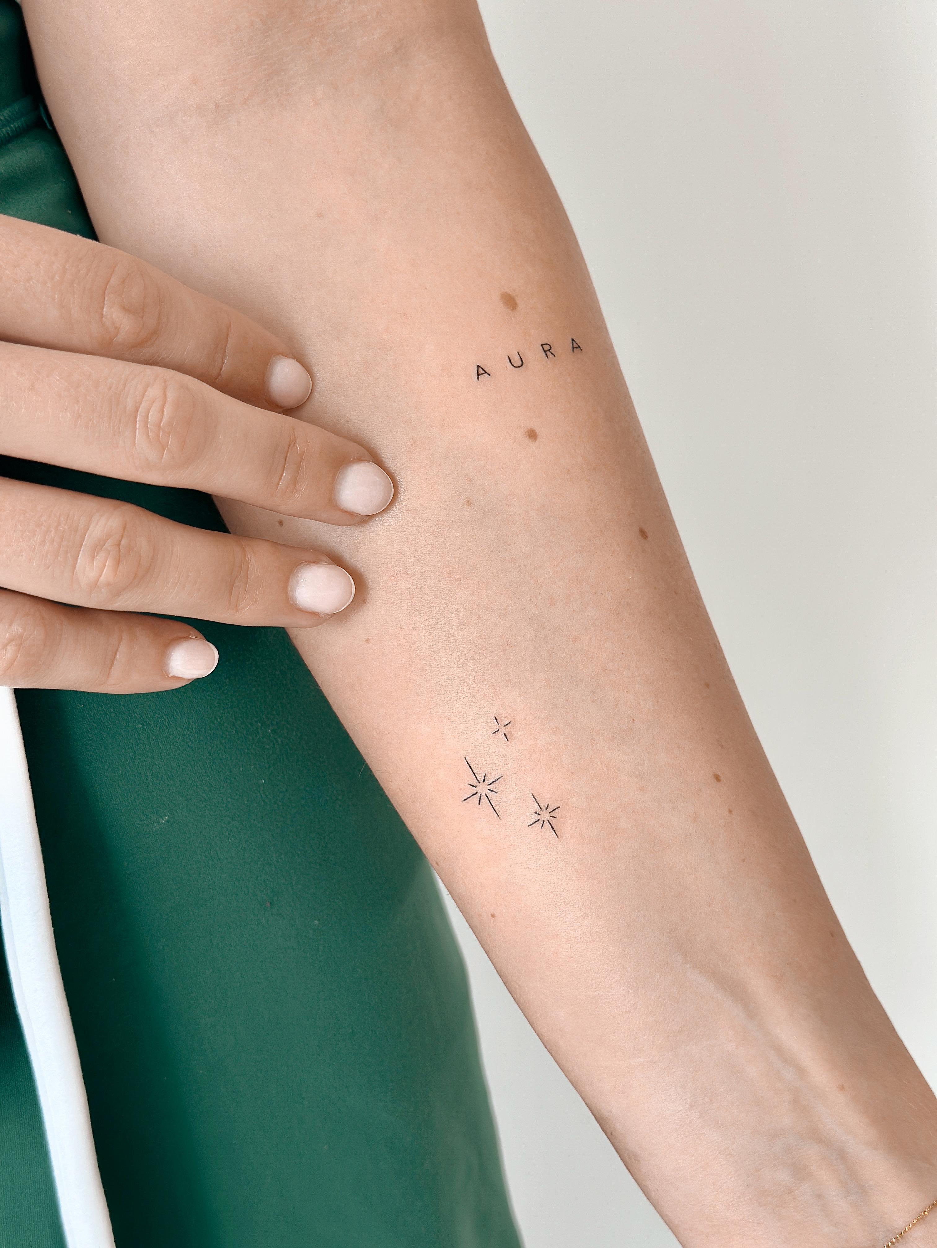 Fine line lettering tattoo reading AURA with small stars